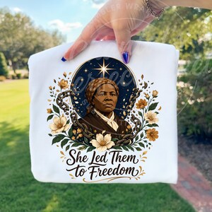 May include: A white t-shirt featuring a graphic of Harriet Tubman centered within a dark blue starry circle. She is surrounded by broken metal chains and light-colored flowers. Below the illustration, the text reads She Led Them to Freedom.