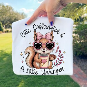 May include: White sweatshirt with a graphic of a cartoon squirrel wearing heart-shaped sunglasses and a pink bow. The squirrel holds a can with a heart and an acorn. Text reads "Cute, Caffeinated & A Little Unhinged."