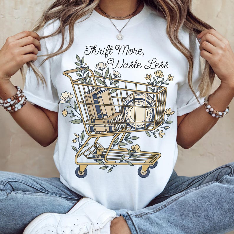 May include: White t-shirt with the text "Thrift More, Waste Less" above a graphic of a shopping cart filled with books and a plate, surrounded by floral elements. The cart is yellow with blue wheels.