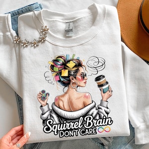 May include: White sweatshirt with a graphic of a woman with colorful hair, sunglasses, and a coffee cup. The text "Squirrel Brain Don't Care" is below the image. A floral necklace and blue jeans are also visible.