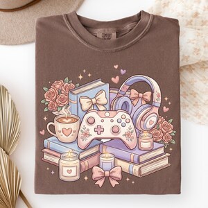May include: Brown t-shirt with a graphic of books, roses, a coffee cup, a game controller, headphones, and candles. The graphic is in pastel colors with hearts and bows. The shirt has the text "Comfort Colors" on the tag.