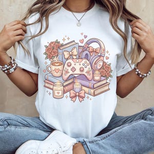 May include: White t-shirt featuring a pastel illustration of books, a coffee cup, a game controller, headphones, candles, and roses. The design includes hearts and bows, creating a cozy and whimsical aesthetic. The shirt is a casual wear item.