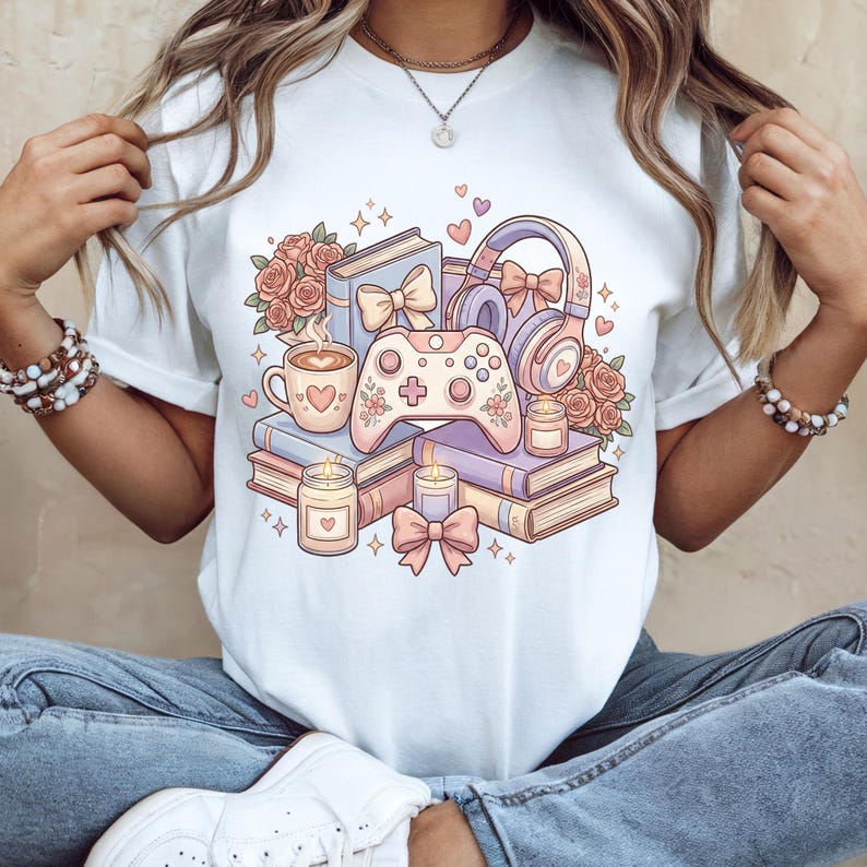 May include: White t-shirt featuring a pastel illustration of books, a coffee cup, a game controller, headphones, candles, and roses. The design includes hearts and bows, creating a cozy and whimsical aesthetic. The shirt is a casual wear item.