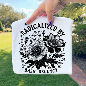 Puede incluir: Sudadera blanca con un diseño floral negro y el texto "RADICALIZED BY BASIC DECENCY". El diseño presenta dos flores grandes y varios elementos florales más pequeños.