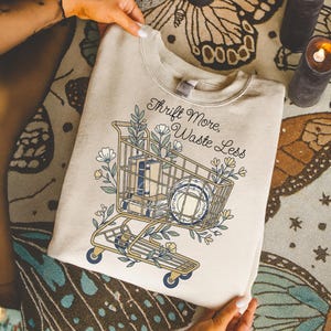 May include: Beige sweatshirt with a shopping cart graphic filled with books and plates, surrounded by floral elements. The text "Thrift More, Waste Less" is arched above the cart. The sweatshirt is held by hands.
