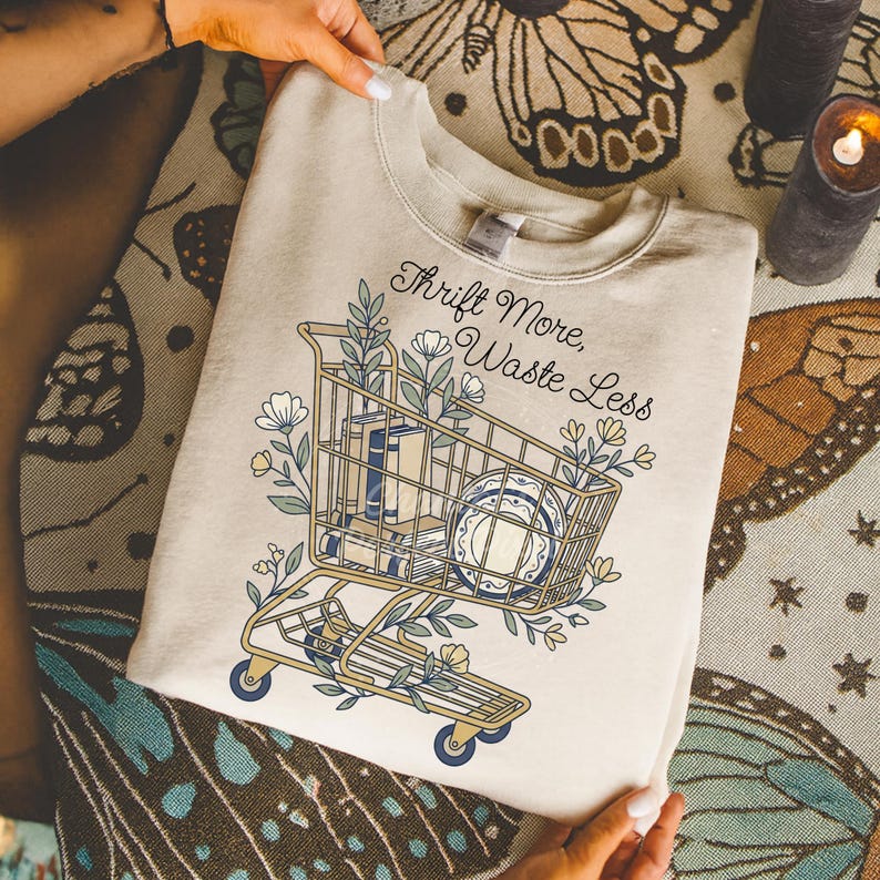 May include: Beige sweatshirt with a shopping cart graphic filled with books and plates, surrounded by floral elements. The text "Thrift More, Waste Less" is arched above the cart. The sweatshirt is held by hands.