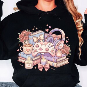 May include: Black hoodie featuring a colorful illustration of a gaming setup with books, a coffee cup, a game controller, headphones, candles, and roses. The design has a pastel color palette with hearts and bows.