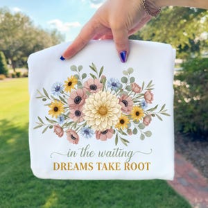 May include: White sweatshirt featuring a floral design with yellow, pink, and blue flowers. The text "in the waiting DREAMS TAKE ROOT" is printed below the floral arrangement. The sweatshirt is held up by a hand.