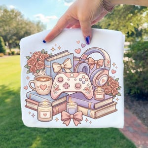 May include: White graphic t-shirt featuring a pastel illustration of books, a coffee cup, a game controller, headphones, roses, candles, and bows. The design has a soft, romantic aesthetic with hearts and floral accents, perfect for a gamer or book lover.