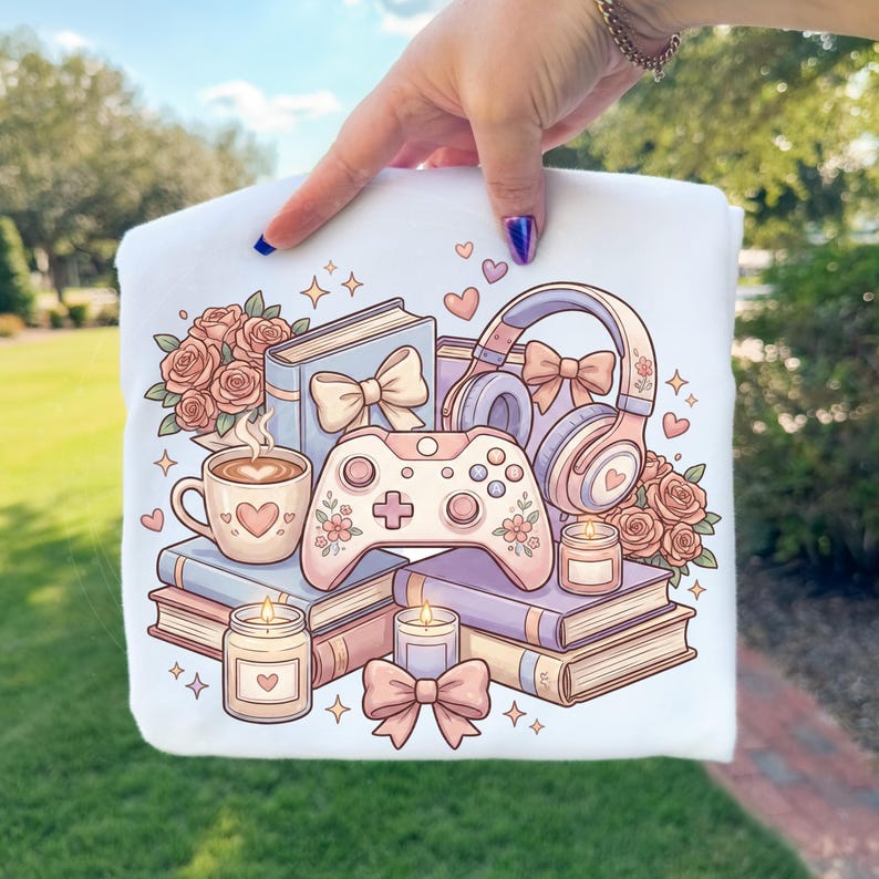 May include: White graphic t-shirt featuring a pastel illustration of books, a coffee cup, a game controller, headphones, roses, candles, and bows. The design has a soft, romantic aesthetic with hearts and floral accents, perfect for a gamer or book lover.