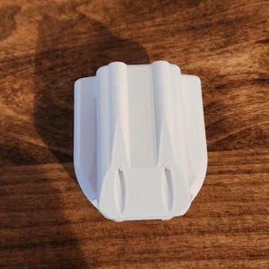 May include: A white, 3D-printed object with a unique, symmetrical design. The object has a rectangular base and a series of vertical, curved ridges. The item is sitting on a wooden surface.