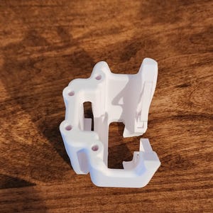 May include: A white 3D-printed object with a complex, geometric design. It features multiple holes and a series of angled surfaces. The object is resting on a wooden surface, showcasing its intricate structure and form.