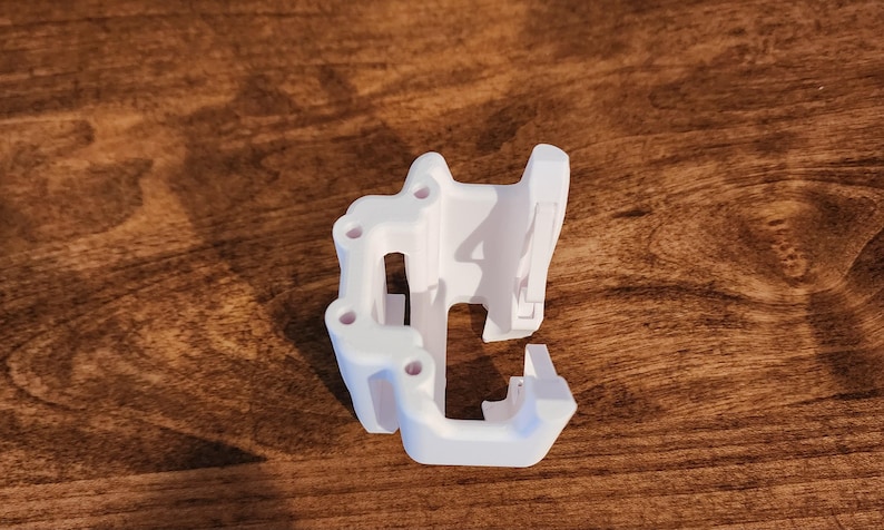 May include: A white 3D-printed object with a complex, geometric design. It features multiple holes and a series of angled surfaces. The object is resting on a wooden surface, showcasing its intricate structure and form.