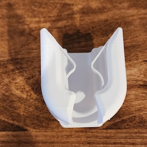May include: A white, 3D-printed object with a U-shaped design. The object has two curved sides that meet at the top, with a central cavity. The item is made of plastic and sits on a wooden surface.