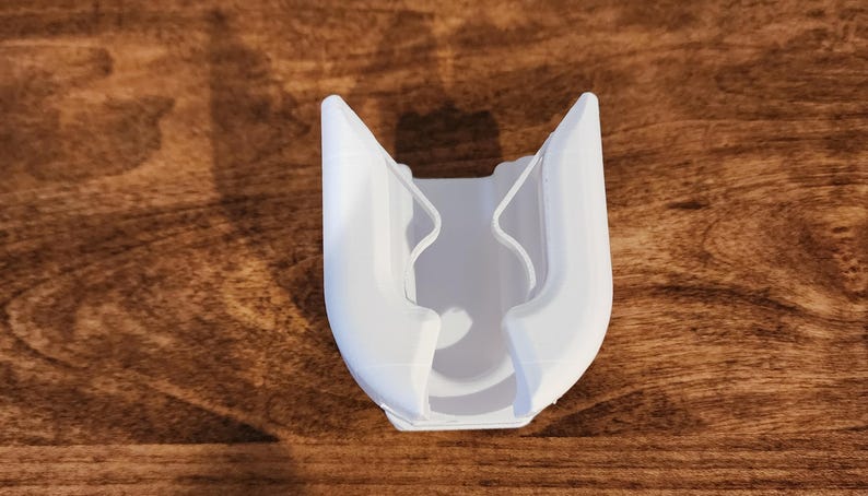 May include: A white, 3D-printed object with a U-shaped design. The object has two curved sides that meet at the top, with a central cavity. The item is made of plastic and sits on a wooden surface.