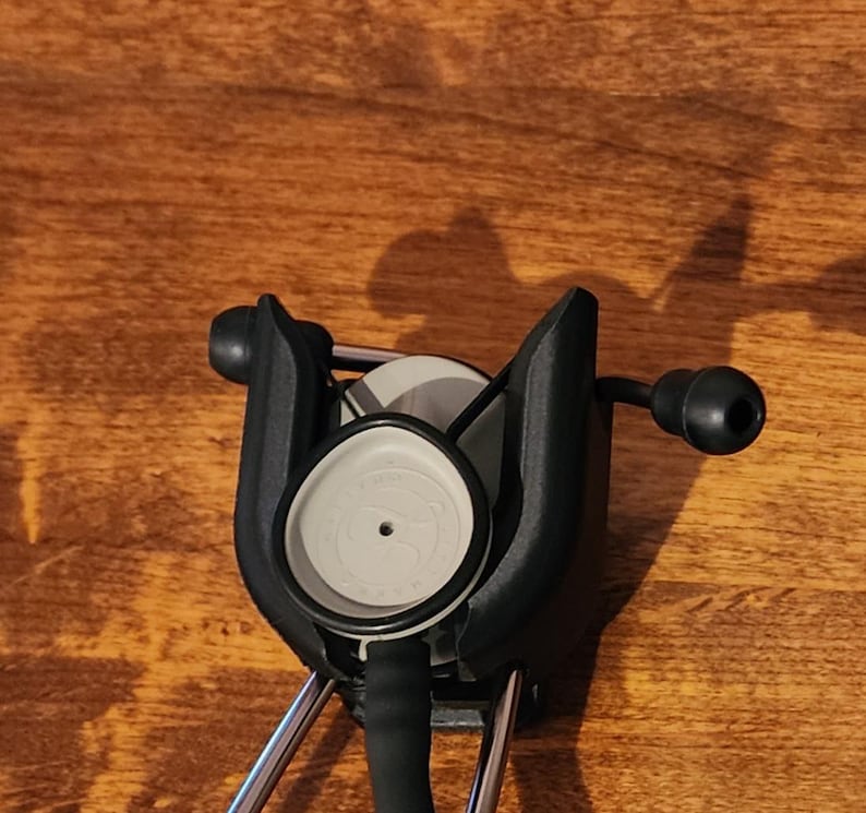 May include: A black and silver stethoscope with a gray diaphragm and black earpieces. The stethoscope is positioned against a wooden surface, with the focus on the chest piece and earpieces.