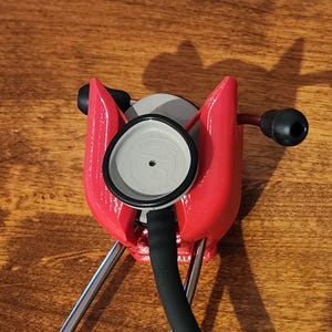 May include: A red stethoscope holder with a stethoscope in place. The holder is made of a bright red material and has a unique shape. The stethoscope has a black tube and ear pieces, and a gray chest piece.