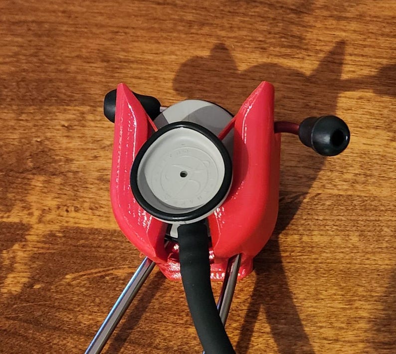 May include: A red stethoscope holder with a stethoscope in place. The holder is made of a bright red material and has a unique shape. The stethoscope has a black tube and ear pieces, and a gray chest piece.