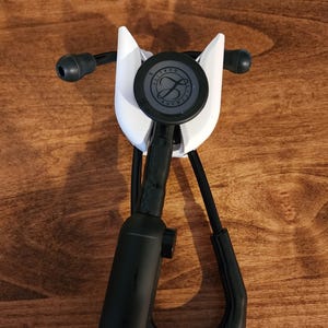 May include: A white stethoscope holder with a black stethoscope. The holder has a curved design and is mounted on a wooden surface. The stethoscope has black tubing and a circular chest piece with a logo.