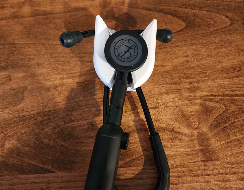 May include: A white stethoscope holder with a black stethoscope. The holder has a curved design and is mounted on a wooden surface. The stethoscope has black tubing and a circular chest piece with a logo.