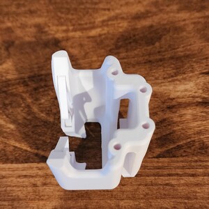 May include: A white, 3D-printed object with a complex, geometric design. It features multiple holes and a series of connected, angular shapes. The object is set against a dark wood grain background.