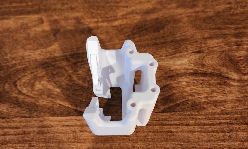 May include: A white, 3D-printed object with a complex, geometric design. It features multiple holes and a series of connected, angular shapes. The object is set against a dark wood grain background.