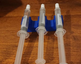 May include: Three clear syringes with white plungers are held in a blue plastic holder. The syringes have labels with text and are filled with a gray liquid. The syringes are on a wooden surface.