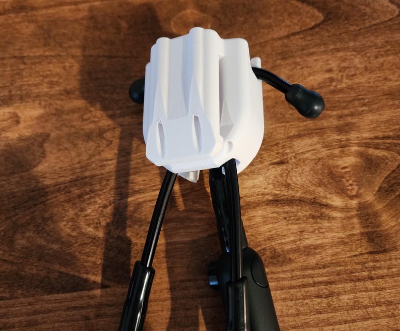 May include: A white plastic device with a rectangular body and two black arms extending downwards. The device has a textured surface and is set against a wooden background. The arms have black, rounded ends.