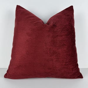 May include: A square, claret-coloured cushion. The cushion has a textured, woven appearance and a soft, plush look. The cushion is a solid colour and is likely used for home decor.