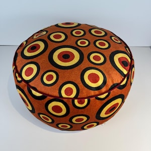 May include: Round orange pouf with a repeating pattern of black, yellow, and red concentric circles. The pouf has a dark red trim around the edge. This decorative pouf can be used as a footrest or extra seating.