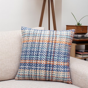 Puede incluir: Cojín decorativo cuadrado con un patrón tejido en tonos azules, naranjas y crema. El cojín está colocado sobre un sofá beige, mostrando su diseño texturizado y paleta de colores. Dimensiones aproximadas: 45 cm x 45 cm.