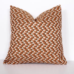 Puede incluir: Cojín decorativo cuadrado con un patrón geométrico de triángulos marrones, blancos y naranjas. El cojín tiene forma cuadrada y está colocado sobre un fondo blanco. Es un artículo de decoración para el hogar.