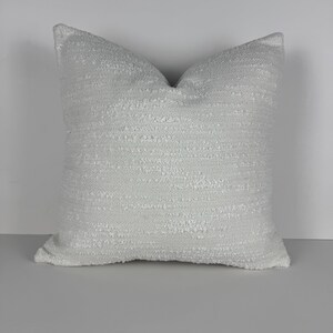May include: A square, textured white pillow. The pillow has a soft, nubby texture and a neutral color, suitable for various decor styles. The pillow is a decorative item for a bed or sofa.