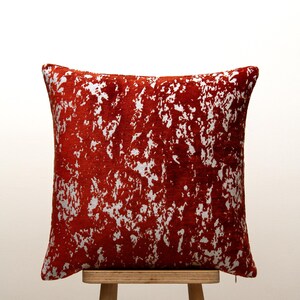 Puede incluir: Un cojín decorativo cuadrado con fondo rojo y un patrón plateado abstracto. El cojín está colocado sobre un taburete de madera. El diseño presenta un aspecto texturizado y moteado. Aproximadamente 40 cm x 40 cm.