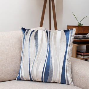 Könnte beinhalten: Quadratisches Zierkissen mit einem wellenförmigen Muster in Blau-, Grau- und Weißtönen. Das Kissen liegt auf einem beigefarbenen Sofa, und das Design erzeugt einen strukturierten, eleganten Look. Die Kanten des Kissens sind dunkelblau. Ca. 40 cm x 40 cm.