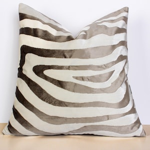 May include: A decorative square pillow with a zebra stripe pattern. The pillow features alternating bands of light beige and dark taupe, creating a bold, eye-catching design. The pillow is made of a soft, textured fabric, perfect for home decor.
