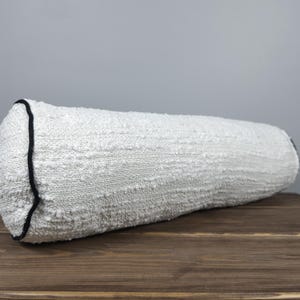 May include: A white bolster cushion with a textured, woven fabric. The ends are finished with a black trim. The cushion is cylindrical and rests on a wooden surface.