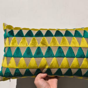Könnte beinhalten: Rechteckiges Zierkissen mit einem geometrischen Muster aus gelben und türkisfarbenen Dreiecken auf beigem Hintergrund. Das Kissen besteht aus einem weichen, strukturierten Stoff. Eine Hand hält das Kissen.