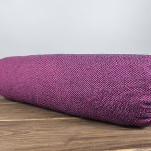 May include: A cylindrical bolster cushion in a deep magenta colour with a textured woven fabric. The cushion is resting on a wooden surface. This item is suitable for yoga, meditation, or as a decorative accent.