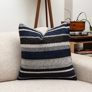 May include: A square decorative pillow with a striped pattern in navy blue, black, gray, and white. The pillow is placed on a beige sofa, offering a cozy and stylish accent for home decor. The pillow's design adds a touch of texture and visual interest.