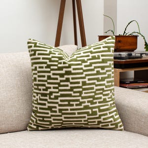 May include: A square decorative pillow with a green and off-white geometric pattern. The pillow is on a beige sofa. The pattern is a series of horizontal and vertical lines, creating a modern design. The pillow adds a pop of color and texture to the living space.