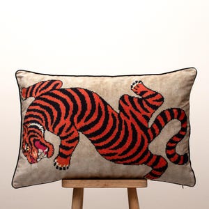 Peut inclure: Coussin rectangulaire décoratif avec un motif de tigre stylisé. Le tigre présente des rayures noires sur un corps rouge, sur un fond beige. Le coussin a une bordure noire et est posé sur un tabouret en bois.