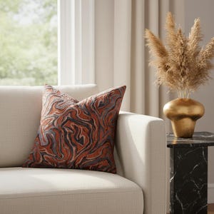 May include: A decorative throw pillow with a swirling pattern in shades of rust, gray, and black rests on a cream-colored sofa. The pillow's design resembles marble or flowing water, adding a touch of elegance to the living space. A gold vase with dried plants sits nearby.