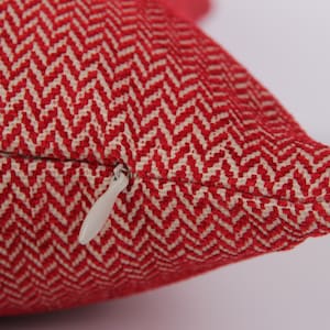 Op de afbeelding: Close-up van een rood en crèmekleurig kussen met een visgraatpatroon. Het kussen heeft een ritssluiting met een witte ritstrekker. De stof is strak geweven en het kussen is waarschijnlijk voor de interieurdecoratie.