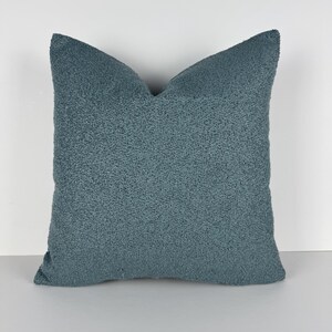 Puede incluir: Un cojín decorativo cuadrado de color verde azulado con una superficie texturizada y nudosa. El cojín tiene un aspecto suave y acogedor y probablemente se utiliza para la decoración del hogar. El cojín es de color liso y tiene un diseño sencillo y moderno.