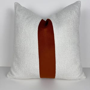May include: A square cushion with a white textured fabric and a vertical stripe of rust-coloured fabric down the centre. The cushion is a neutral colour and would complement a variety of home decor styles.
