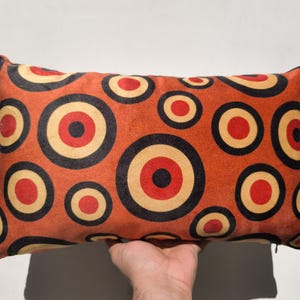 Op de afbeelding: Rechthoekig sierkussen met een herhalend patroon van concentrische cirkels in rood, zwart en beige op een oranje achtergrond. Het kussen wordt door een hand omhoog gehouden. Het heeft een ritssluiting.