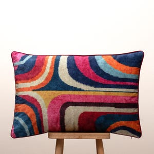 Könnte beinhalten: Ein rechteckiges Zierkissen mit einem lebendigen, von Retro inspirierten Design. Das Kissen zeigt ein Muster aus geschwungenen Streifen in Blau-, Rosa-, Orange-, Gelb- und Weißtönen. Die Kanten sind mit einem dunkelroten Rand versehen, der einen Hauch von Eleganz verleiht.