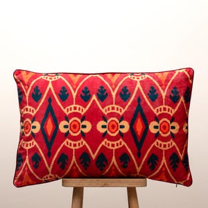 May include: A rectangular cushion in red, featuring a geometric design in orange, teal, and cream. It has a black border and is placed on a wooden stool. The pattern incorporates diamond and floral motifs.