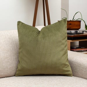 May include: A square, olive green corduroy pillow with vertical ribbed texture. The pillow is placed on a light beige sofa, offering a cozy and textured accent for home decor. The pillow's color adds a touch of nature-inspired elegance.